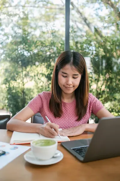Bir kafede dizüstü bilgisayar kullanırken deftere yazan genç bir kadın. Parlak doğal ışık ve dışarıdaki yeşillik.