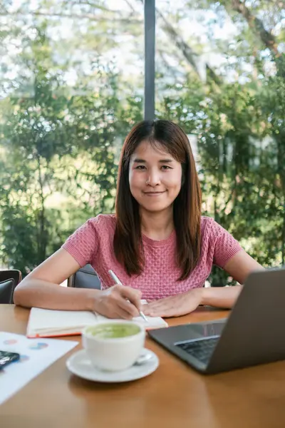 Pembe tişörtlü genç bir kadın bir kafede dizüstü bilgisayarda çalışıyor. Etrafı yeşilliklerle çevrili. Matcha latte içiyor..