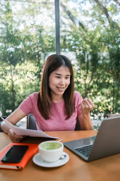 Bir kafede dizüstü bilgisayarda çalışan gülümseyen bir kadın. Etrafı yeşilliklerle çevrili, elinde kahve ve belgelerle..
