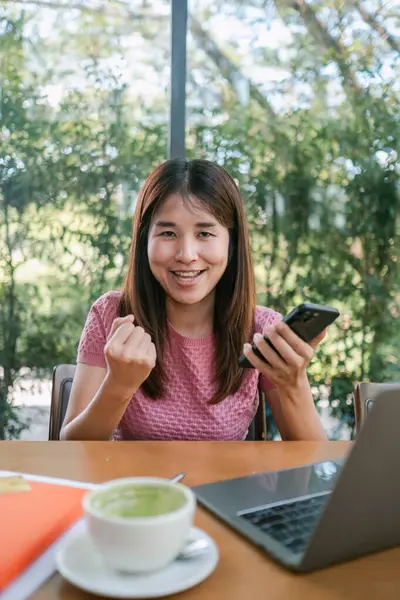 Bir kafede gülümseyen kadın, başarıyı akıllı telefon ve dizüstü bilgisayarla kutluyor, yeşilliklerle çevrili..