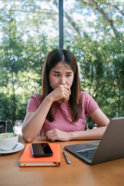 Bir kafede genç bir kadın, laptopuna odaklanmış, etrafı yeşilliklerle çevrili, masada bir not defteri ve kahveyle.