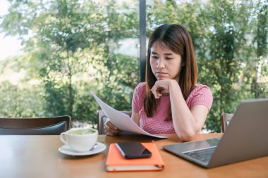 Pembe tişörtlü bir kadın, pencerenin dışındaki yeşilliklerle çevrili bir laptopla ve kahveyle masa başında dokümanları inceliyor..