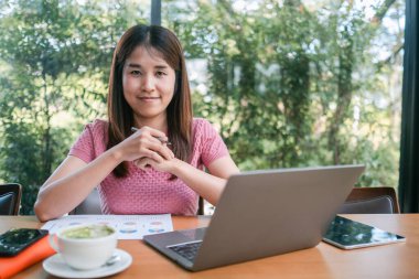 Pembe tişörtlü genç bir kadın bir kafede dizüstü bilgisayarla çalışıyor, etrafı yeşilliklerle çevrili, elinde bir fincan kahveyle..