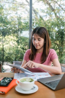 Genç bir kadın, modern bir çalışma ortamını gösteren dizüstü bilgisayar, kahve ve belgelerle çevrili, parlak bir kafede tablet kullanıyor..