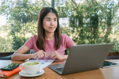 Yeşilliklerle çevrili bir kafede dizüstü bilgisayar kullanan odaklanmış genç bir kadın. Masasında kahve ve belgeler var..