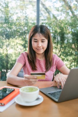 Genç bir kadın yeşil çay lattesiyle rahat bir kafede otururken bilgisayarını ve kredi kartını online alışveriş için kullanıyor..