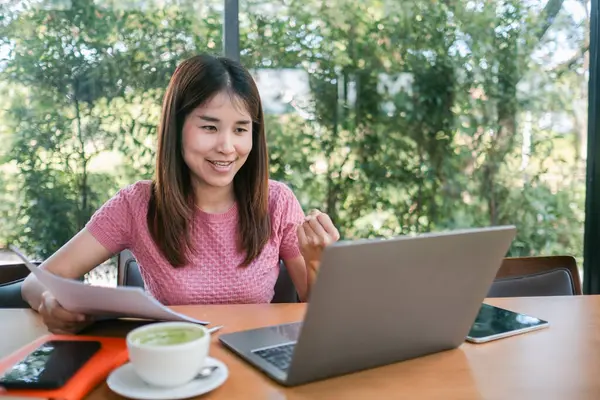 Pembe gömlekli neşeli genç bir kadın, dışarıda yemyeşil yemyeşil bir kafede laptopuyla çalışırken başarısını kutluyor..