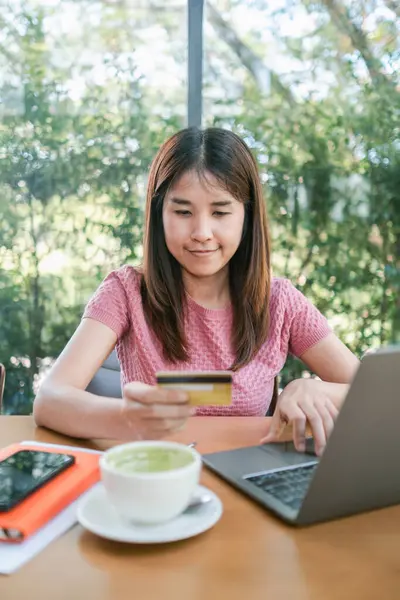 Genç bir kadın yeşil çay lattesiyle rahat bir kafede otururken bilgisayarını ve kredi kartını online alışveriş için kullanıyor..