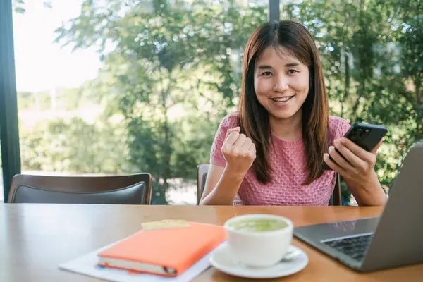 Elinde akıllı bir telefonla gülümseyen genç bir kadın, modern bir kafede dizüstü bilgisayarı ve defteri olan bir masada oturuyor.