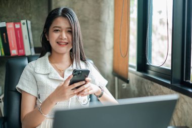 Akıllı telefonlu kendinden emin genç bir kadın, modern ofis ortamında gülümsüyor, pencerenin kenarında oturuyor..
