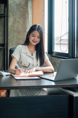 Genç bir kadın masada oturuyor, dizüstü bilgisayarı açık bir şekilde yazıyor, parlak ve modern bir ofis ortamında..