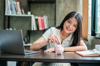Gülümseyen kadın, kumbaraya para koyuyor. Birikimleri ve finansal planlamaları simgeliyor..
