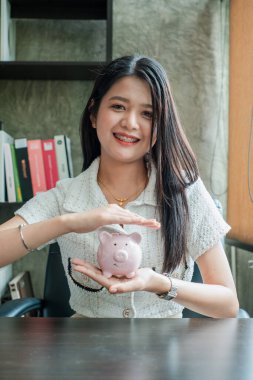 Ofiste elinde domuz kumbarası olan neşeli bir kadın. Tasarruf, finans ve gelecek planlarını temsil ediyor..