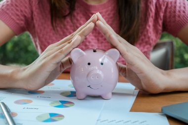 Finansal belgeler ve renkli çizelgelerle çevrili pembe bir kumbarayı koruyan bir kadının yakın çekimi..