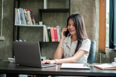 Günümüz ofis ortamında dizüstü bilgisayar ve akıllı telefon kullanan kendine güvenen genç bir kadın..