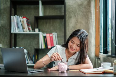 Gülümseyen kadın, ofis masasındaki kumbarada para biriktirip mali planlama ve bütçeyi sembolize ediyor..