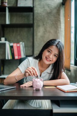 Gülümseyen kadın ofis masasındaki domuz kumbarasına para koyuyor. Tasarruf ve finansal planlamayı simgeliyor..