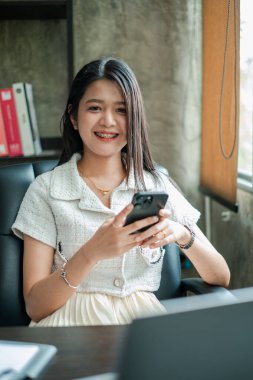 Akıllı telefonlu genç bir kadın, modern ofis ortamında gülümsüyor, profesyonelliği ve bağlantıyı aktarıyor..