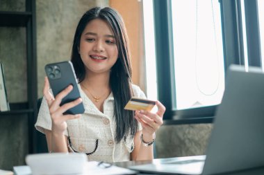 Evde online alışveriş için akıllı telefon ve kredi kartı kullanan gülümseyen kadın, pencerenin yanında dizüstü bilgisayar başında oturuyor..