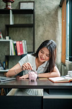 Gülümseyen kadın, modern ofis masasındaki kumbarada kitap ve dizüstü bilgisayarla para biriktiriyor..