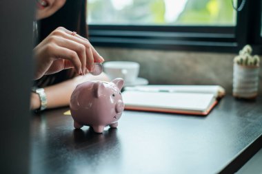 Birikimleri ve finansal planlamaları sembolize eden, masanın üzerindeki pembe kumbaraya bozuk para sokan bir kadının yakın plan çekimi..