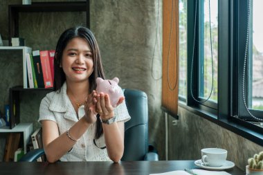 Sırıtkan kadın elinde bir kumbara, para ve tasarruf simgesi, masada kitaplar ve kahveyle..