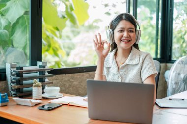 Laptop ve kulaklık kullanan gülümseyen bir kadın, dışarısı yeşilliklerle kaplı, modern bir ofiste uzaktan çalışıyor..
