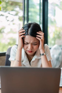 Kadın, ofisteki dizüstü bilgisayarda çalışırken strese giriyor. Pencereden doğal ışık ve yeşillik görünüyor..