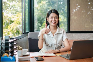 Ofiste gülümseyen genç bir kadın başparmak kaldırıyor, dizüstü bilgisayarla ve belgelerle masada oturuyor, pozitiflik ve başarı sergiliyor..