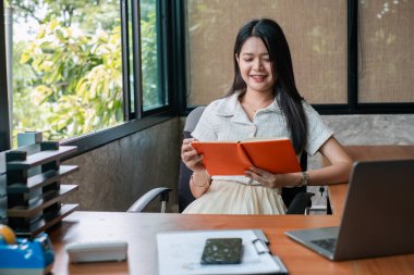 Modern ofis ortamında tablet okuyan genç bir kadın. Oda dışarısı doğal ışık ve yeşilliklerle dolu..