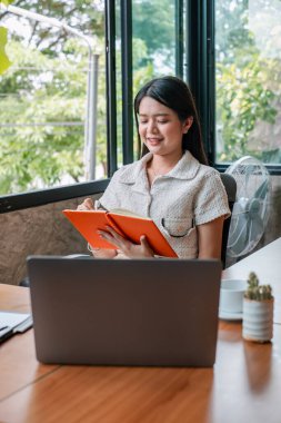 Genç bir kadın dizüstü bilgisayarda çalışıyor ve dışarıda büyük pencereleri ve yeşillikleri olan parlak ve modern bir ofiste notlar alıyor..