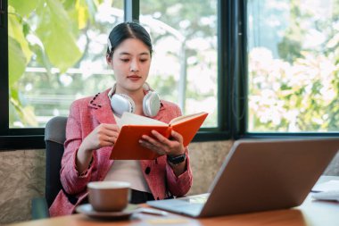 Modern bir ofisteki genç bir kadın, masasında bir kitap okuyor, dizüstü bilgisayarı ve kulaklıkları var, doğal ışık ile çevrili..