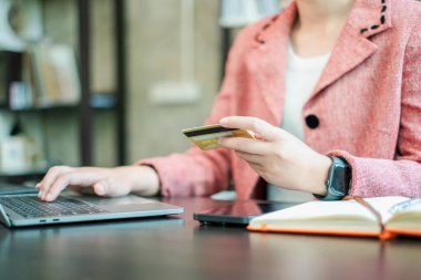 Pembe ceketli biri dizüstü bilgisayar kullanıyor ve elinde kredi kartı tutuyor. İnternet üzerinden alışveriş ve iş işlemlerini temsil ediyor..
