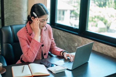 Pembe ceketli profesyonel kadın, ofis masasında dizüstü bilgisayarı, defteri ve hesap makinesiyle çalışmaya odaklanmış..