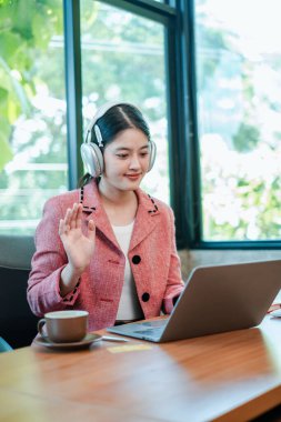 Pembe ceketli genç bir kadın, parlak ve modern bir ofiste video görüşmesi için dizüstü bilgisayar ve kulaklık kullanıyor..