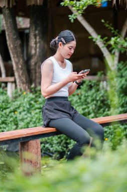 Yeşil bir parktaki ahşap bir bankta oturan günlük kıyafetli genç bir kadın akıllı telefonuna odaklanmış..