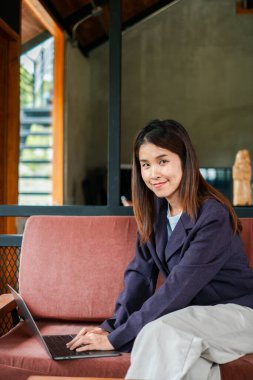 Genç bir kadın, bir kanepede rahat bir şekilde oturur. Ahşap aksanlı, modern ve iyi aydınlatılmış bir iç dizüstü bilgisayar kullanır..