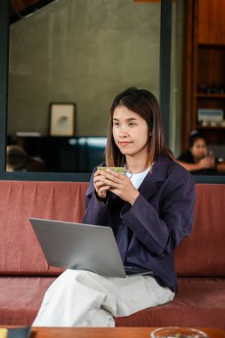 Kafede elinde kupa olan bir kadın laptopuyla çalışıyor, rahat ve üretken bir atmosfer yaratıyor..