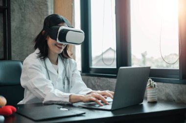 VR kulaklık takan gülümseyen kadın doktor, parlak bir tıp ofisinde dizüstü bilgisayarla yazı yazmak, sağlık hizmetlerinde teknolojiyi kucaklamak..