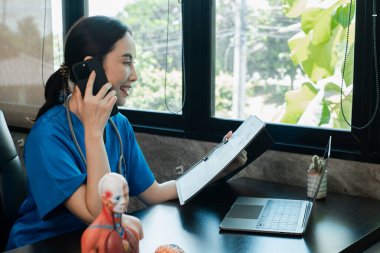 Mavi önlüklü bir sağlık görevlisi bir masada oturuyor, akıllı telefon kullanıyor ve belgeleri inceliyor. Bir dizüstü bilgisayar ve anatomik model görünür.