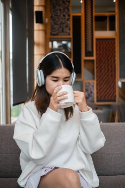Genç bir kadın, şık, rahat bir oturma odasında kulaklıkla müzik dinlerken bir fincan kahveden zevk alır..