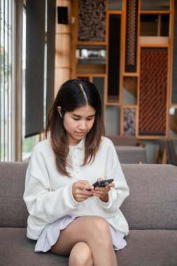 Genç bir kadın, şık dekorlu modern bir oturma odasında akıllı telefonuna odaklanmış bir şekilde koltuğa rahatça oturur..