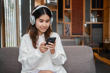 Modern bir kafede akıllı telefon kullanan, kulaklık takan genç bir kadın. Rahat ve teknoloji meraklısı atmosfer.