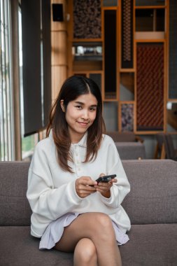 Şık, modern bir kafe içinde, koltukta otururken akıllı telefon kullanan gülümseyen genç bir kadın..