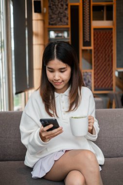 Genç bir kadın kahvesini içiyor ve şık, modern bir kafe ortamında akıllı telefonunu karıştırıyor..