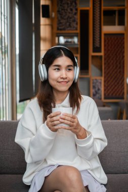 Genç bir kadın, modern bir oturma odasında, kulaklıklar ve bir fincan kahveyle huzurlu bir anın tadını çıkarıyor..