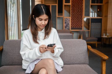 Beyaz kazaklı genç bir kadın şık ve modern bir kafe ortamında akıllı telefonunu kullanıyor..