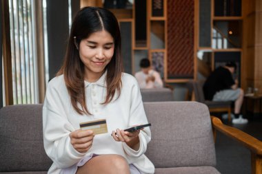 Genç bir kadın rahat bir kafede otururken online alışveriş için akıllı telefonunu ve kredi kartını kullanıyor..