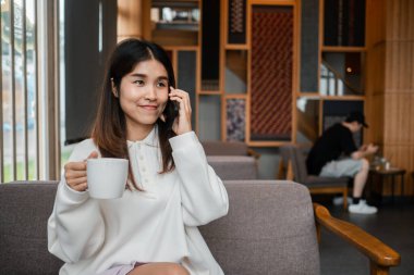 Süslü bir kafede elinde kahve fincanı tutarken telefonda konuşan beyaz kazaklı genç bir kadın..