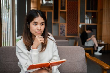 Modern bir kafede elinde bir defter ve kalem tutan genç bir kadın, derin düşüncelere dalmış. Sıcak, ahşap dekorlu davetkar bir atmosfer..